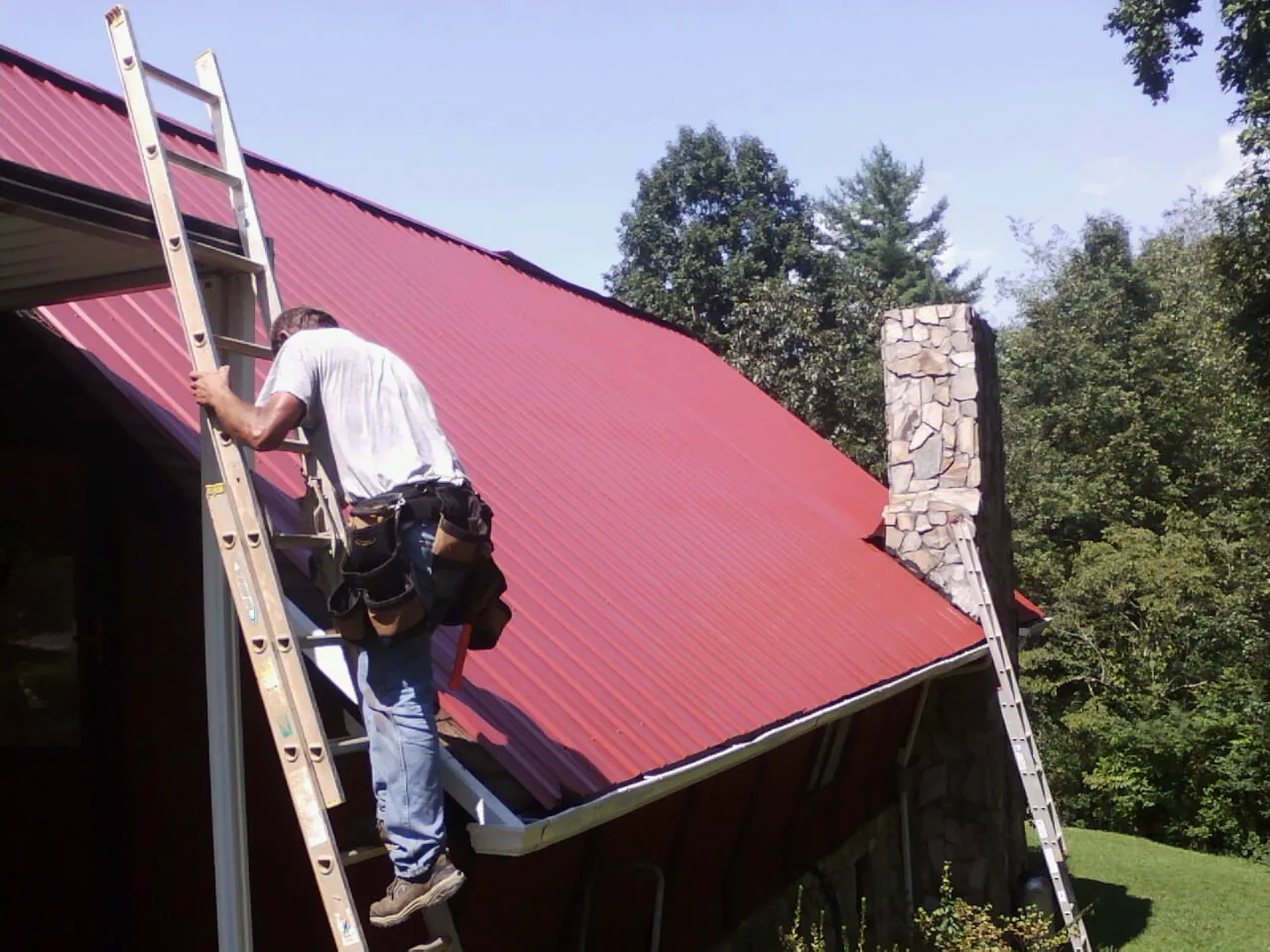 Roofer on a ladder beside a red metal roof and stone chimney — Soffit & Fascia Repair by Roofing Brothers in Star